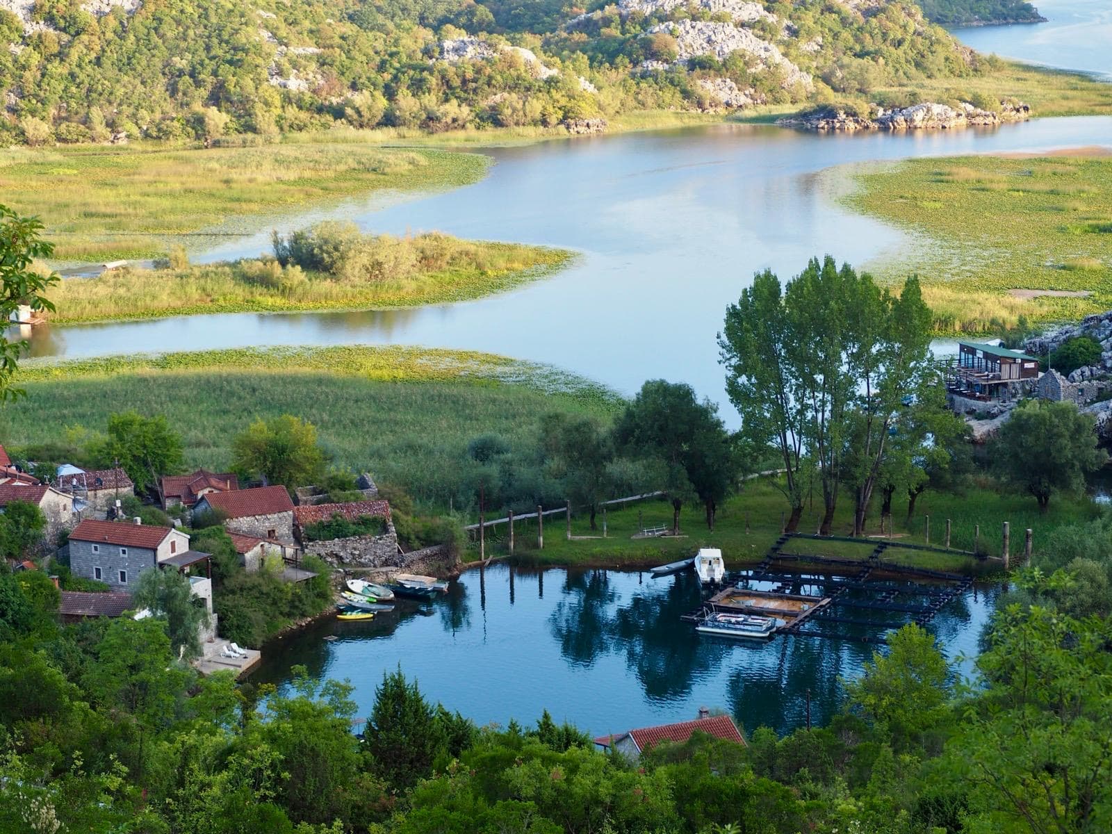 Aerial view of Karuc fishing village showing traditional stone houses, small harbor with boats, and Lake Skadar's pristine waters surrounded by lush green hills